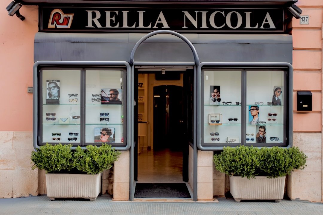 Vetrine dell'ottica con insegna Rella Nicola al civico 137, occhiali Prada e Dior in esposizione e vasi di bosso all'ingresso