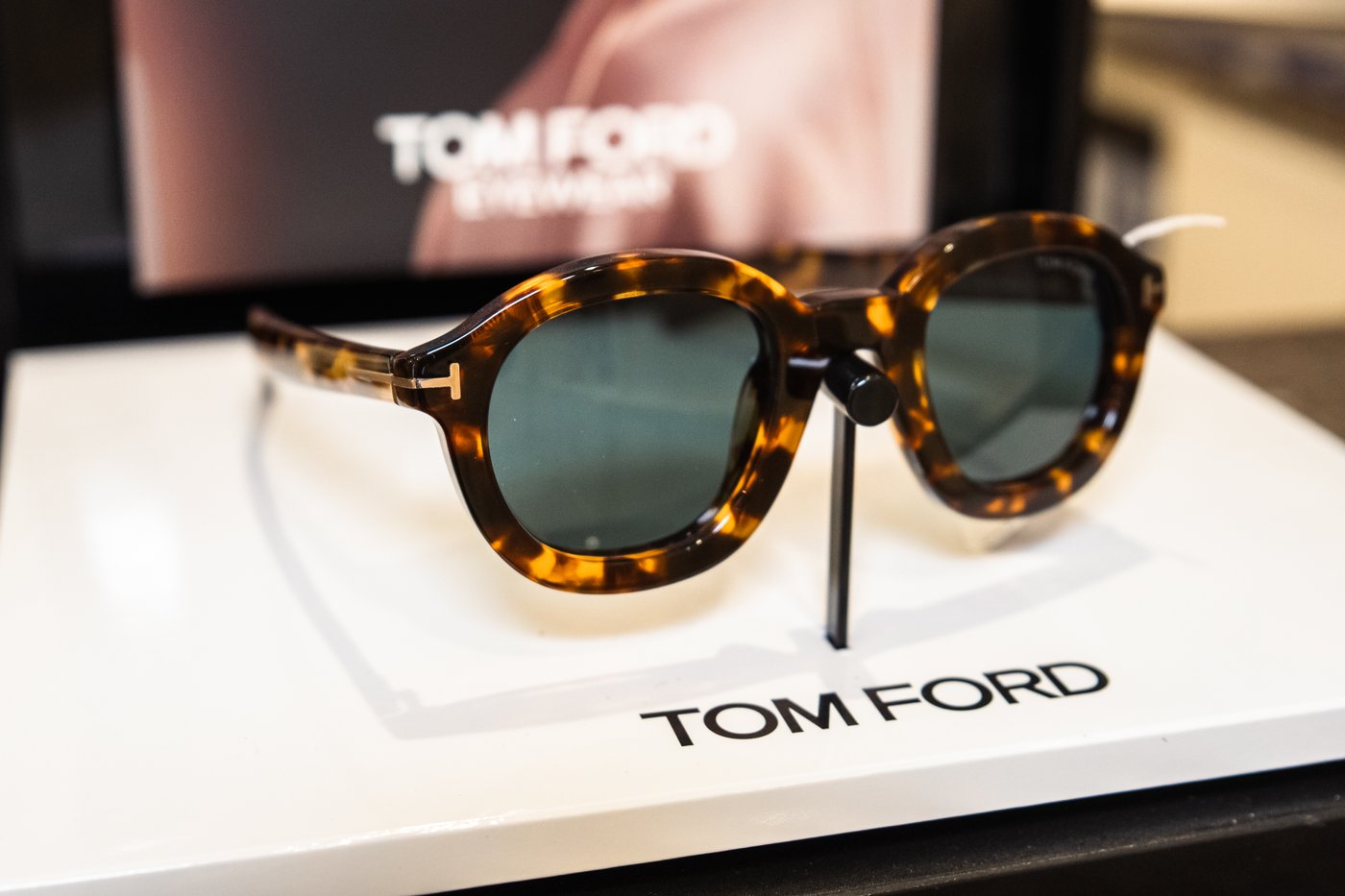 Occhiali da sole Tom Ford in acetato tartarugato con lenti verdi su espositore bianco con logo, alle spalle pannello Eyewear