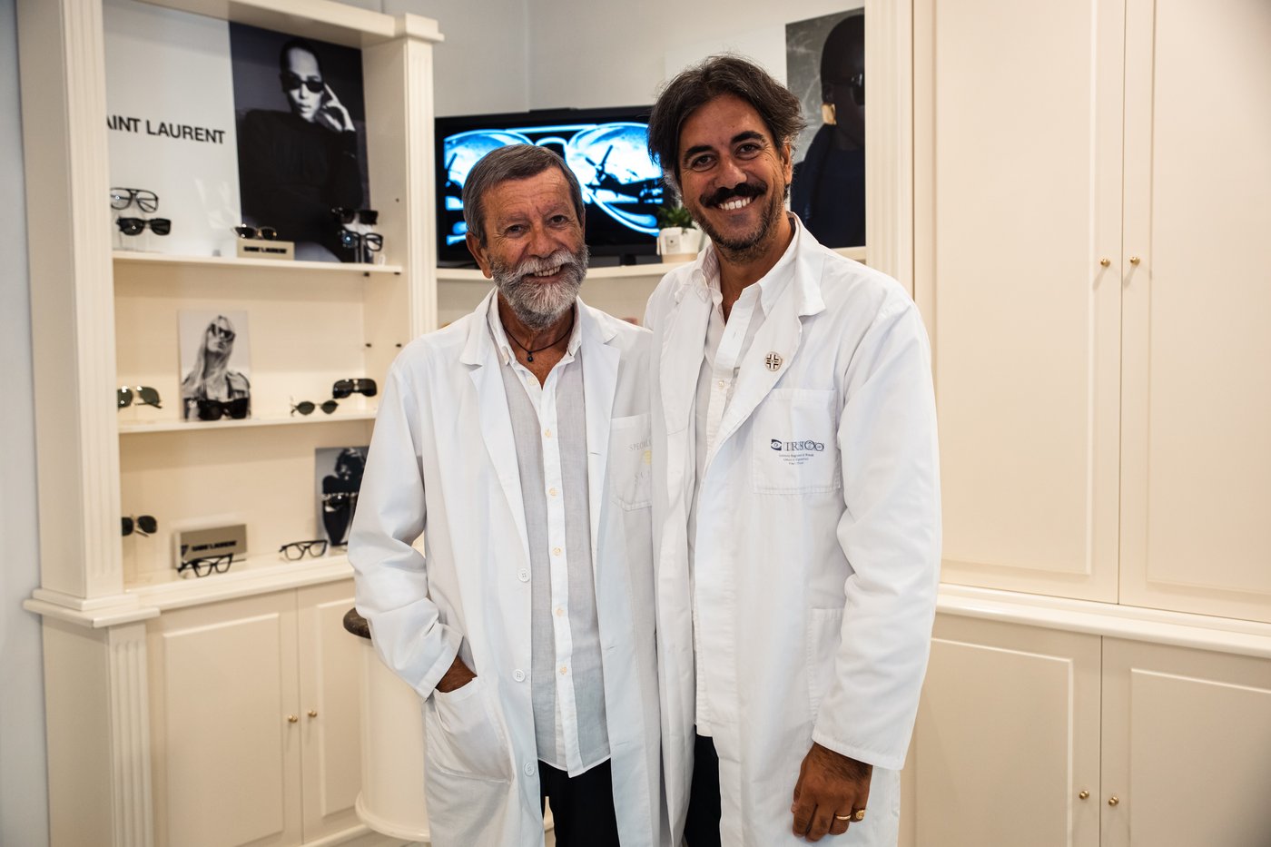 Due ottici in camice bianco sorridono davanti a scaffali color crema con occhiali da sole e cartello Saint Laurent
